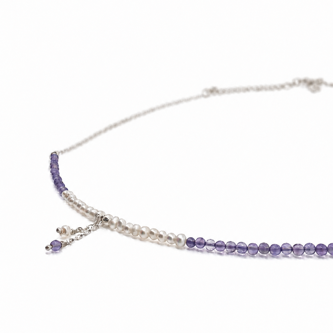 A delicate silver-toned necklace featuring a strand of white pearls and a strand of purple beads, with a small dangling charm, shown on a plain white background.