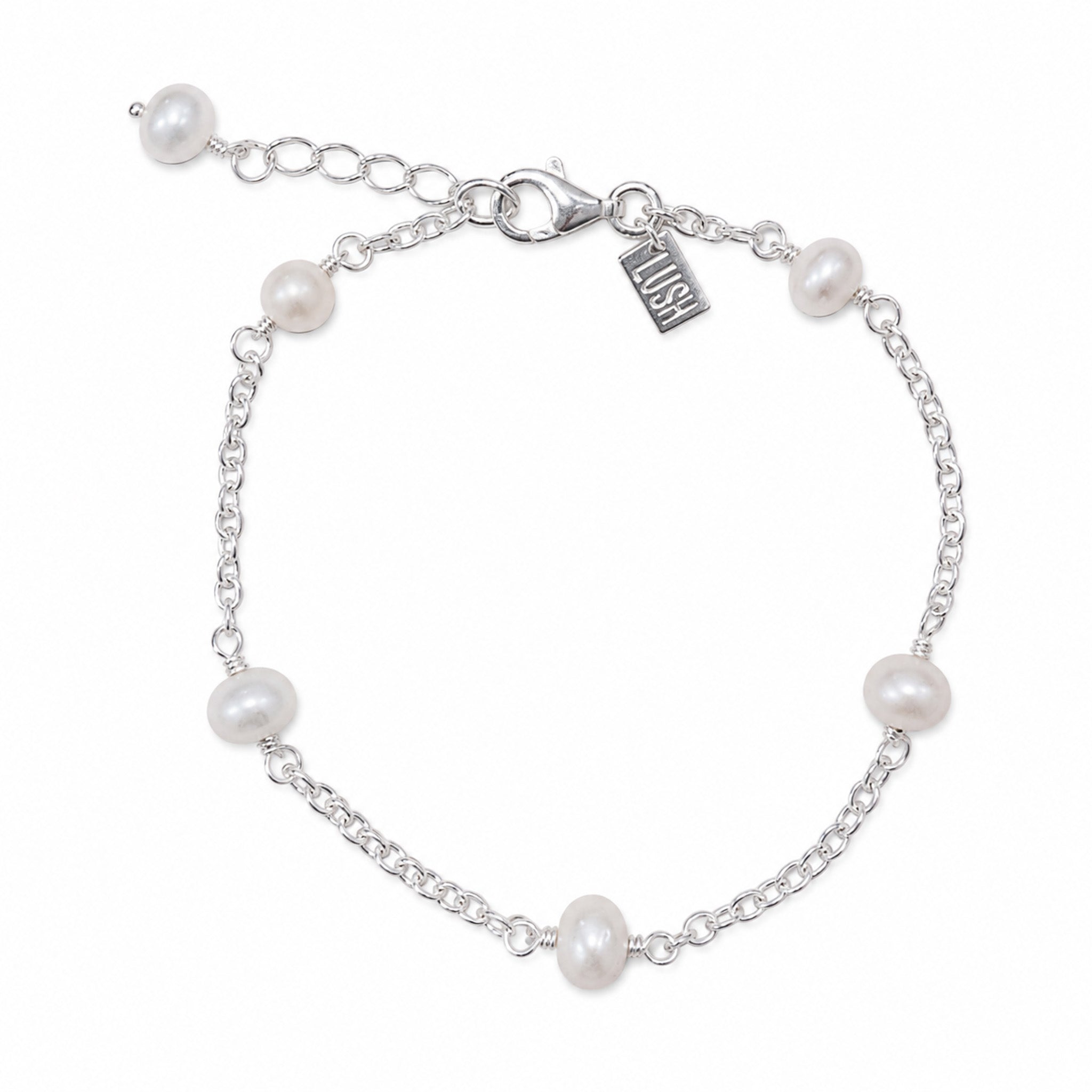 A silver chain bracelet with six white freshwater pearls spaced evenly along the chain, featuring a lobster clasp, extender chain, and a small engraved brand tag that reads 'LUSH'.