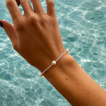 A delicate silver chain bracelet with three white freshwater pearls, worn on a woman's wrist against a sunny, sun-dappled swimming pool background.